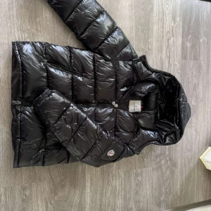 Svart Moncler pufferjacka med huva - Säljer en svart, glansig pufferjacka från Moncler med huva och dragkedja framtill. Jackan har flera fickor med dragkedja och klassisk Moncler-logga på ärmen. Insidan har ett coolt serietidningsmönster. Perfekt för kalla dagar och riktigt snygg streetstil.