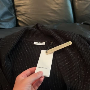 Svart kofta från Lindbergh med knappar - Svart ribbad kofta från Lindbergh med snygg krage och knäppning framtill. Långärmad modell i mjukt material med diskreta ränder och återvunnen bomull. Perfekt för lager-på-lager och chill stil.