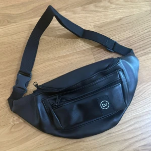 Svart midjeväska - Helt Ny!!! Snygg svart magväska från DV i skinnimitation med justerbart band och två dragkedjefack framtill. Perfekt för att bära mobilen, plånboken och andra småprylar. Enkel och stilren design med DV-logga på framsidan.
