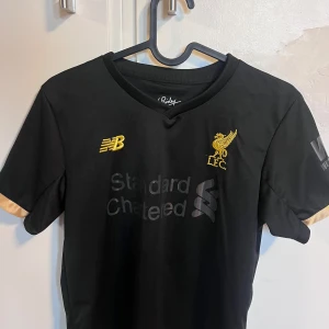 Liverpool Salah tröja svart/guld New Balance - Liverpool FC fotbollströja i svart med guldiga detaljer från New Balance. Tryck med M. Salah och nummer 11 på ryggen, klubbmärke och sponsorlogga på bröstet. Korta ärmar med guldfärgade muddar. Material i lätt och ventilerande polyester.