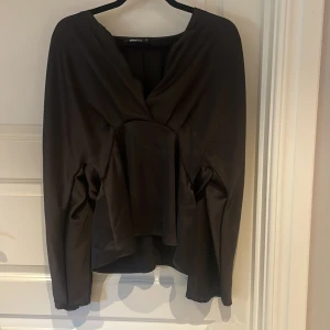 Svart omlottopp från Gina Tricot - Säljer en svart omlottopp från Gina Tricot i storlek 36. Toppen har lång ärm, djup v-ringning och snyggt draperad omlottdesign framtill. Materialet är mjukt och har en lätt glansig finish som ger en elegant känsla.