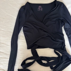Svart omlottopp från Crivit - Svart långärmad omlottopp från Crivit med knytband i midjan. Toppens v-ringning och croppad passform ger en snygg och modern look. Perfekt att styla med höga jeans eller kjol för en trendig outfit.