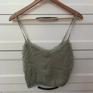 Ljusgrön stickad linnetopp FB Sister - Supersöt ljusgrön stickad linnetopp från FB Sister med smala axelband, spetskant och liten rosett fram. Toppen är croppad och har en mjuk, ribbad struktur med diskreta hålmönster. Perfekt för varma dagar eller lager på lager-stil.