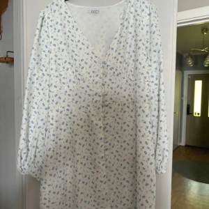Vit blommig klänning från NA-KD - Supersöt vit klänning från NA-KD med små blå blommor, v-ringning och knappar framtill. Långa puffärmar med resår vid ärmslut. Perfekt för vår och sommar, luftig och lätt i bomull.