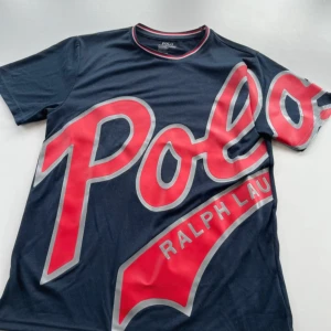 Polo Ralph Lauren t-shirt med stor logga - Två snygga t-shirts från Polo Ralph Lauren med stor, fet logga över hela framsidan. En är marinblå med röd och vit text, den andra är vit med svart text. Båda har rund hals och korta ärmar. Perfekt statement-plagg för dig som gillar streetstyle.