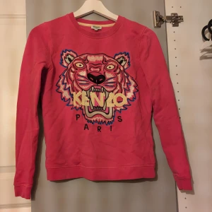 Rosa Kenzo sweatshirt med tigertryck - Säljer en rosa sweatshirt från Kenzo med ikoniskt broderat tigerhuvud och Kenzo Paris-text på bröstet. Tröjan har rund hals och långa ärmar. Perfekt statement piece för dig som gillar färg och coola prints. Storlek xs men passar även xxs