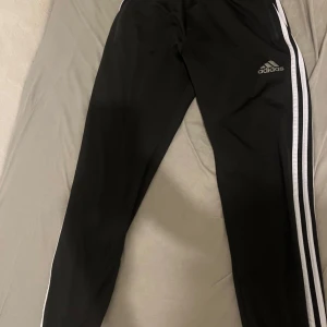 Svart Adidas träningsset med vita ränder - Säljer ett svart Adidas träningsset med klassiska vita ränder längs ärmar och ben. Jackan har dragkedja, ståkrage och logga på bröstet samt fickor med dragkedja. Byxorna har smal passform och Adidas-logga på låret. Perfekt för dig som gillar sportig stil.