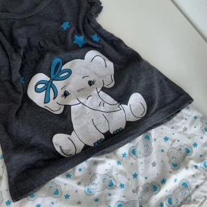 Pyjamas sätt elefantmotiv - Supersöt mörkgrå ärmlös topp med ett stort tryck av en tecknad elefant med blå rosett och blå stjärnor framtill. Toppen är i mjuk bomull och har en avslappnad passform. Shortsen kom med tröjan som ett sätt. Elefantmotivet ger en gullig och lekfull vibe.