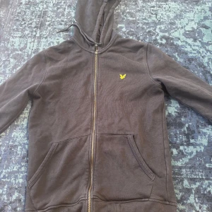 Svart hoodie från Lyle & Scott - Svart hoodie från Lyle & Scott med dragkedja framtill och klassisk gul logga på bröstet. Tröjan har huva med snörning, två fickor och ribbade muddar. Perfekt för chill dagar eller när du vill ha en enkel och stilren look.