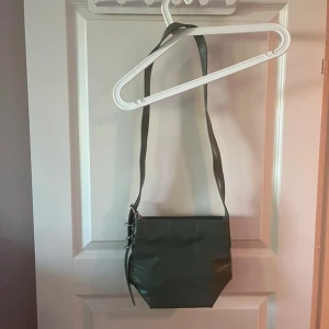 Grön axelväska i skinn - Snygg grön axelväska med stilren design och justerbart axelband. Väskan har en unik, kantig form och detaljer i silverfärgad metall. Perfekt för dig som vill ha en enkel men trendig accessoar till din outfit.