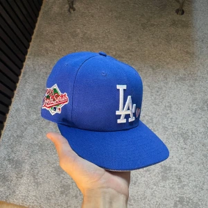 LA keps New Era - Snygg blå keps från New Era med LA Dodgers-logga framtill och broderad basebollhjärta. På sidan finns 1988 World Series-märke och bak står det 'Love & Hustle = Game' broderat. Klassisk rak skärm och MLB-logga bak. Perfekt för dig som älskar baseball och streetstyle.