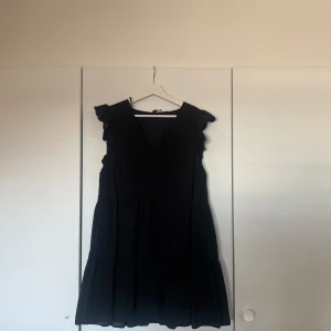 Svart volangklänning med v-ringning - Svart kortklänning med v-ringad hals och volangdetaljer vid axlarna. Klänningen har en luftig passform och är ärmlös, perfekt för en stilren look. Tillverkad i ett lätt material som ger ett snyggt fall.