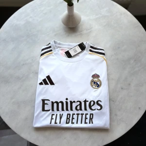 Real Madrid Adidas matchtröja S - Säljer en officiell Real Madrid fotbollströja från Adidas i storlek S. Tröjan är vit med svarta detaljer, gula inslag och klubbmärke på bröstet. Klassiska Adidas-ränder på axlarna och 'Emirates Fly Better' tryckt framtill. Tillverkad i lätt och ventilerande material.