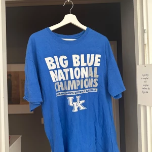 Blå Kentucky Wildcats t-shirt - Säljer en blå t-shirt med stort tryck 'Big Blue National Champions' och Kentucky Wildcats-logga framtill. Perfekt för dig som gillar basket och vill visa ditt stöd för UK. Klassisk passform och rund hals.