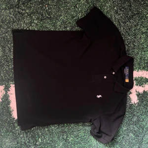 Svart pikétröja från Ralph Lauren - Klassisk svart pikétröja från Ralph Lauren med vit broderad logga på bröstet. Tröjan har korta ärmar, krage och två knappar framtill. Perfekt för en clean och stilren look, funkar året runt och är lätt att matcha med allt. OBS: inte org1inal