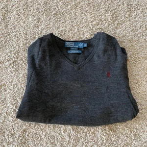 Mörkgrå v-ringad tröja Polo Ralph Lauren - Snygg mörkgrå v-ringad tröja från Polo Ralph Lauren i mjuk merinoull. Klassisk passform med långa ärmar och diskret röd logga broderad på bröstet. Perfekt att slänga på över en skjorta för en clean och stilren look.