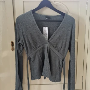 Balsam green Tie Top från Gina Tricot - Grön omlottopp från Gina Tricot med knytband i midjan och lång ärm. Toppen är oanvänd med lappen kvar. Orginal pris 299