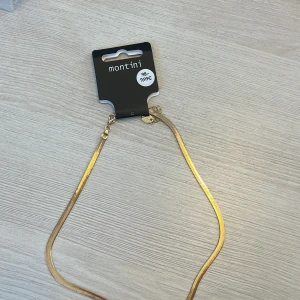 Guldigt halsband från Montini - Stilrent och platt guldigt halsband från Montini. Kedjan har en elegant, slät design som ger en modern känsla och passar perfekt till olika looks. Justerbar längd med karbinhake för enkel användning. Helt oanvänt