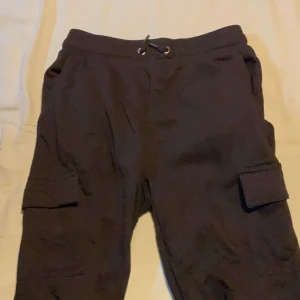 Svarta cargopants från R-COVER - Svarta cargopants från R-COVER med snörning i midjan och stora fickor på sidorna. Byxorna har en loose passform och är tillverkade i mjukt material som känns skönt mot huden. Perfekta för en avslappnad och cool stil.