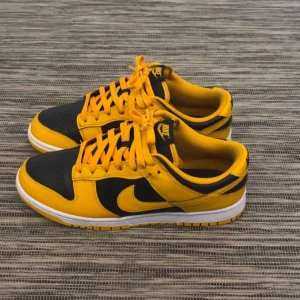 Nike Dunk Low - Nike Dunk Low sneakers i svart och gul med klassisk vit sula. Skorna har snörning, rund tå och är tillverkade i skinn och syntet. Perfekt för dig som vill sticka ut med en färgstark och ikonisk modell.