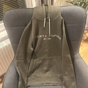 Olivgrön hoodie Tommy Hilfiger (PRISET KAN DISKUTERAS VID SNABB AFFÄR) - Snygg olivgrön hoodie från Tommy Hilfiger med broderad logga och text på bröstet. Tröjan har huva med vita snören och en stor magficka. Perfekt för en chill och avslappnad stil. Materialet känns mjukt och skönt mot huden. Den är dessutom knappt använd och i nyskick.