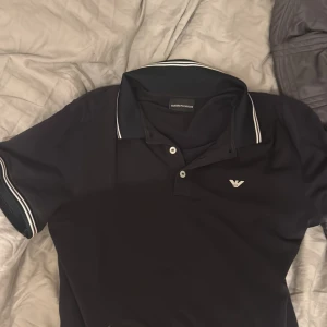 Svart pikétröja från Emporio Armani - Snygg svart pikétröja från Emporio Armani med vit logga på bröstet och vita ränder på krage och ärmslut. Klassisk polokrage med två knappar. Perfekt för en stilren och sportig look. 150kr