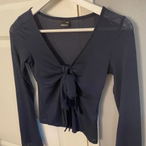 Mörkblå knytblus från Gina Tricot - Supersnygg mörkblå långärmad topp från Gina Tricot i storlek XXS. Modellen har djup v-ringning och en stor knytning framtill som ger en trendig look. Tunn och mjuk i materialet, perfekt att styla med jeans eller kjol.