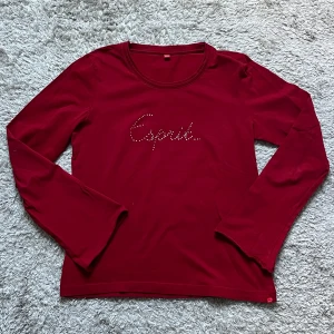 Röd långärmad tröja från Esprit - Snygg röd långärmad tröja från Esprit med glittrande stenar som bildar loggan på bröstet. Har aningen vidare öppning kring handlederna. Tröjan har rund halsringning och är gjord i mjuk bomull, perfekt för dig som gillar enkel men stilren design. 