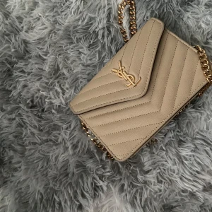 Beige quiltad axelväska från Saint Laurent - Säljer en beige axelväska från Saint Laurent med klassisk YSL-logga i guld framtill. Väskan är quiltad med snygga diagonala sömmar och har en kedjerem i guld. Utsidan är i skinn och insidan har en vinröd dragkedjeficka. Perfekt för dig som gillar lyxiga detaljer. Kan diskutera pris.