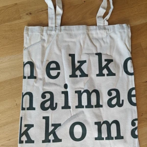 Beige tygkasse med svart text - Säljer en helt oanvänd shoppingväska / tygkasse i bomull med stora bokstäver tryckta på framsidan. Väskan har handtag och är perfekt för att bära böcker, mat eller annat du behöver under dagen. Enkel och stilren design som passar till det mesta. Perfekt på stranden. Tjockt, canvasliknande tyg och verkar vara va väldigt hög kvalitet. Ca 45x38 cm.