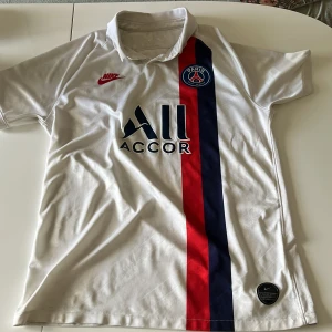 PSG vit matchtröja Nike M - Säljer en vit Paris Saint-Germain fotbollströja från Nike i storlek M. Tröjan har klassisk krage, korta ärmar och Dri-FIT-material. Framsidan har en röd och marinblå vertikal rand, PSG-logga och Nike Swoosh i rött. Perfekt för dig som älskar fotboll och PSG.