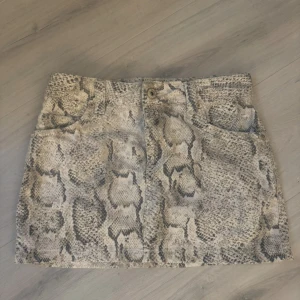 Beige kortkjol med ormmönster - Snygg kortkjol i beige och grå toner med coolt ormmönster. Kjolen har klassisk jeansmodell med fickor fram och bak samt hällor för bälte. Perfekt för dig som vill sticka ut med en trendig look.