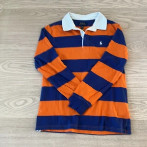 Randig pikétröja från Polo Ralph Lauren. Ny pris 2195 - Snygg långärmad pikétröja från Polo Ralph Lauren med breda orange och marinblå ränder, klassisk vit krage och broderad logga på bröstet. Perfekt för dig som gillar preppy stil och vill sticka ut med färg. Tillverkad i mjuk bomull.