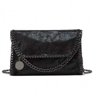 Svart axelväska med kedjedetaljer - Snygg svart axelväska i syntetmaterial med coola kedjedetaljer runt kanterna och som axelrem. Väskan har ett kuvertformat lock och en rund metallbricka som dekoration. Perfekt för dig som gillar en edgy och trendig stil. Liknar Stella mccartney