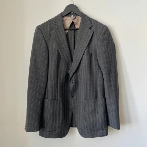 Tommy Hilfiger grå kavaj, storlek 50 - Klassisk tvåknäppt kavaj från Tommy Hilfiger, tillverkad i exklusiv virgin wool. Tidlös design med strukturerad passform och högkvalitativt material.  Skick: Bra begagnat skick. Inga fläckar, hål eller revor. Se bilder för egen bedömning.  Storlek: 50 Material: 100% virgin wool  Mått: Längd: 79 cm Bröstvidd: 53 cm Axelbredd: 45 cm Ärmlängd: 65 cm