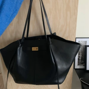 Svart shoppingväska i skinn med gulddetalj - Stor svart shoppingväska i slätt skinn med långa handtag och snygg guldfärgad spänndetalj framtill. Väskan har rymligt foder och innerficka med dragkedja. Perfekt för dig som vill ha plats för allt du behöver under dagen. Lite små defekter på handtaget annars i fint skick! Köpt för 399kr