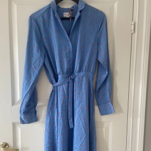 Blå mönstrad klänning Holly & White - Supersöt blå klänning från Holly & White med små röda prickar, knappar framtill och matchande bälte i midjan. Långärmad och med klassisk krage, perfekt för dig som gillar färg och lekfulla detaljer. Knälång🌸använt ett fåtal gånger, gott som ny!