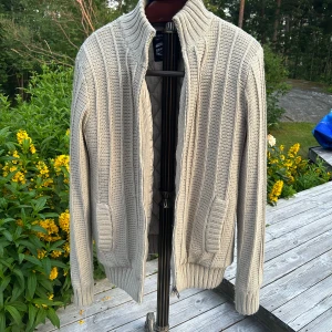 Beige stickad kofta från Angelo Litrico - Snygg beige stickad kofta från Angelo Litrico med ribbade detaljer och dragkedja framtill. Koftan har hög krage, två fickor och quiltat innerfoder för extra värme. Perfekt för lager-på-lager under kyliga dagar.