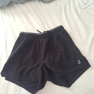 Svarta träningsshorts från SOC - Svarta träningsshorts från SOC med elastisk midja och snörning. Lätta och luftiga med innerbyxa för extra komfort. Perfekta för löpning eller gym, tillverkade i syntetmaterial som andas och håller dig torr.