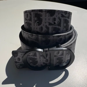 Svart och gråt mönstrat Dior bälte - Snyggt bälte från Dior i svart och grått med ikoniskt CD-spänne i matt svart metall. Bältet har ett grafiskt mönster med Dior-logga över hela bandet och är tillverkat i skinn. Perfekt accessoar för att lyfta din outfit med en lyxig touch.