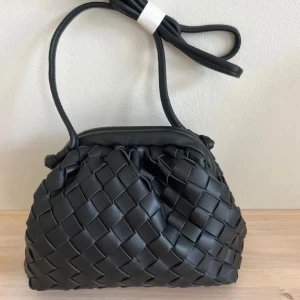 Svart flätad handväska i skinn - Snygg svart handväska med flätad design och axelrem. Väskan har en mjuk, rundad form och är tillverkad i skinn. Perfekt för dig som gillar stilrena accessoarer med en twist. Helt ny 