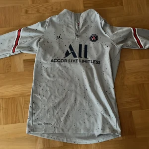 PSG långärmad träningströja Jordan - Snygg PSG träningströja från Jordan i ljusgrått med svarta stänk, klubbmärke på bröstet och dragkedja vid halsen. Röda och svarta detaljer på ärmarna. Tillverkad i funktionsmaterial, perfekt för fotbollsträning eller streetwear.
