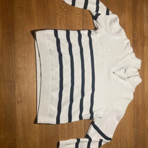 Vit och marinblå randig tröja Lindex - Vit stickad tröja från Lindex med marinblå ränder och half zip-dragkedja vid halsen. Tillverkad i ekologisk bomull, långärmad och med klassisk ribbad mudd. Perfekt för dig som gillar en clean och sportig look.