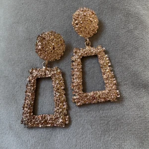Glittriga guldiga statementörhängen - Säljer ett par stora, glittriga örhängen i guld med cool struktur. De har rund platta upptill och rektangulär, öppen form nedtill. Perfekta för dig som vill sticka ut och addera extra bling till din look.