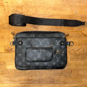 Louis Vuitton svart axelväska - Snygg svart axelväska från Louis Vuitton med klassiskt monogrammönster. Väskan har justerbar axelrem med broderad logga, silverfärgade metalldetaljer och en praktisk ytterficka med dragkedja framtill. Tillverkad i skinn med exklusiv känsla.