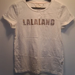 Vit t-shirt LALALAND Holly & Whyte - Vit t-shirt från Holly & Whyte by Lindex med metallicfärgad LALALAND-text på bröstet. Klassisk rund halsringning och korta ärmar. Tillverkad i mjuk bomull, perfekt för en chill och stilren look.