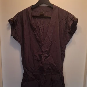 Mörklila kort byxdress Gina Tricot - Byxdress med shorts från Gina Tricot i mörkgrå nyans. Modellen har v-ringning, knappar framtill och korta ärmar med snygga spänndetaljer. Resår i midjan ger en skön passform. Perfekt för dig som gillar en avslappnad laidback stil.