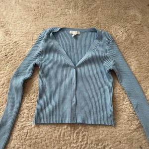 Ljusblå ribbad kofta med v-ringning - Säljer en ljusblå ribbad kofta med v-ringning och långa ärmar. Koftan har knappar framtill och är i ett stretchigt, stickat material som sitter snyggt på kroppen. Perfekt att slänga över en topp eller klänning för en chill look.