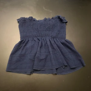 Mörkblå smockad tubtopp - Supersöt mörkblå tubtopp med smockad överdel och volangkant upptill. Toppen har en luftig, lätt utsvängd nederdel och är axelbandslös. Perfekt för varma dagar när du vill ha något bekvämt och stilrent.