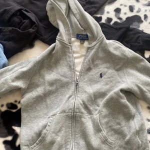 Grå hoodie från Polo Ralph Lauren - Snygg grå hoodie från Polo Ralph Lauren med dragkedja och klassisk blå logga broderad på bröstet. Tröjan har huva, känguruficka och är tillverkad i mjuk bomullsmix. Perfekt för chill dagar eller när du vill ha en clean och enkel look.